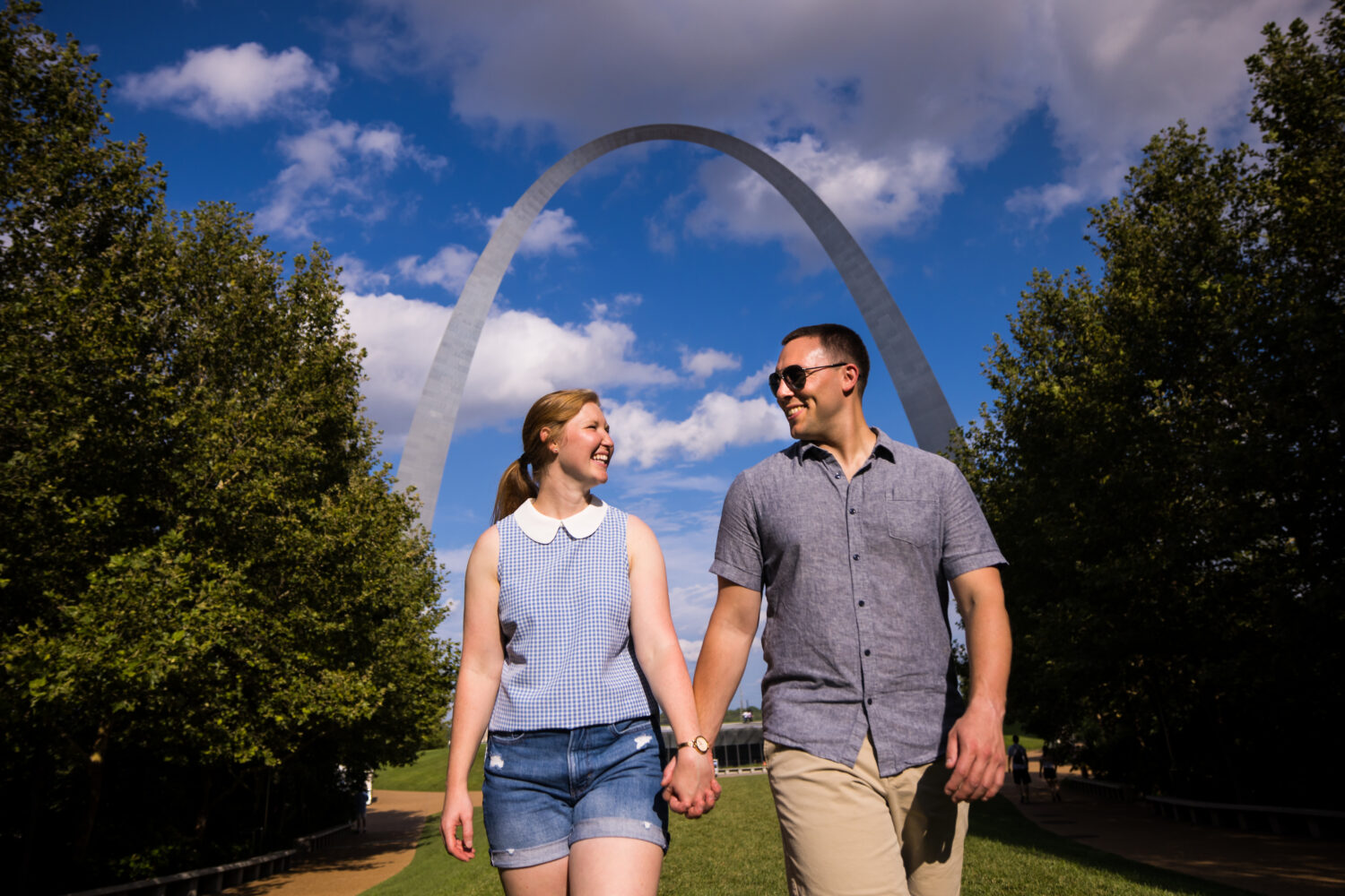 Creative Proposal Photographer, lisa rhinehart, captures this unique, vibrant, fun image of this couple in st louis Missouri with the arch behind them as they walk away from it holding hands and smiling at one another
