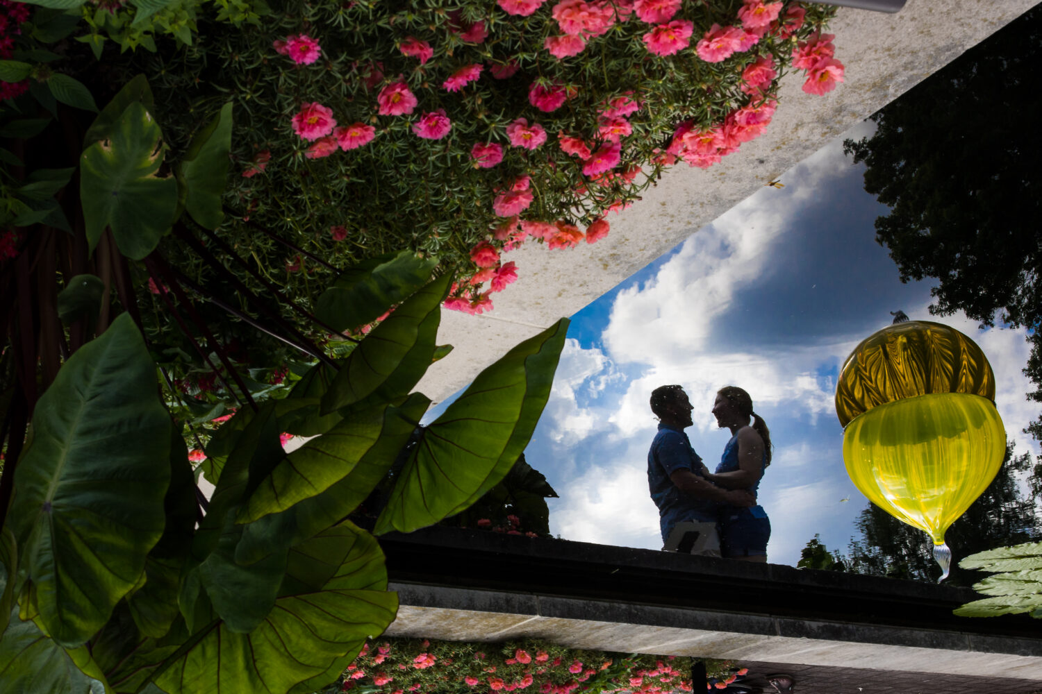 Creative Proposal Photographer, rhinehart photographer, captures this creative, fun, unique image through a reflection in the water of the couple hugging and facing each other surrounded by vibrant colored flowers and plants