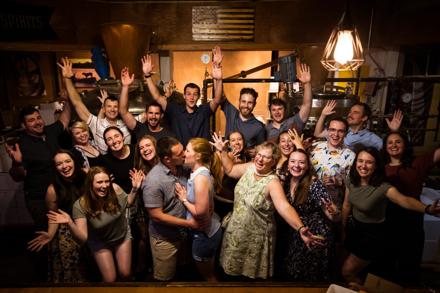 lisa rhinehart, captures this fun image of the couple standing in the middle kissing as they are surrounded by their friends and family at their surprise engagement party after the surprise proposal 