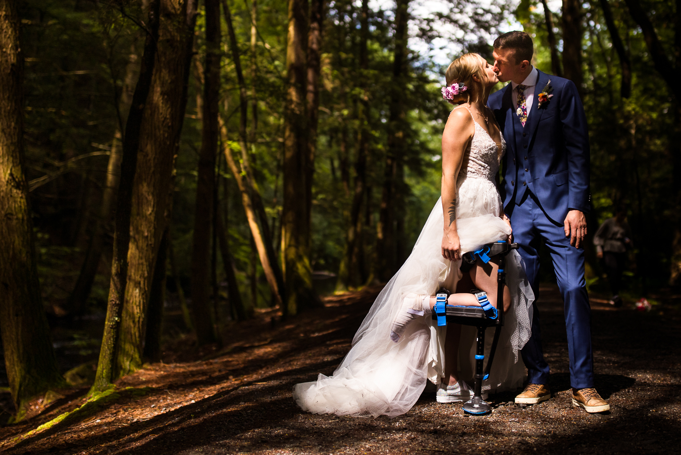 Micro wedding photographer, lisa rhinehart, captures this bride and groom as they kiss one another underneath the trees on a hiking trail showing off the bride's shattered foot from a biking accident