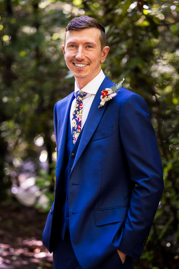 micro wedding photographer, lisa rhinehart, captures this traditional image of the groom smiling at the camera in his vibrant blue tux and floral tie inside of the woods on the hiking trail