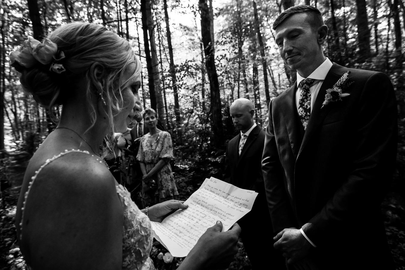 black and white image of the bride holding a piece of paper reading her vows to her groom surrounded by family and friends captured by micro wedding photographer, lisa rhinehart