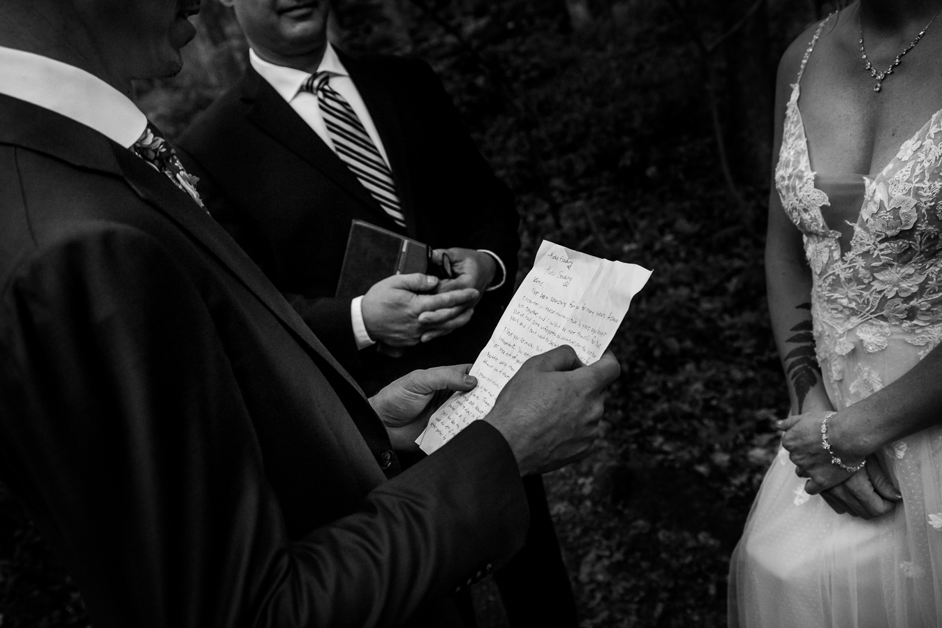 black and white image of the groom holding a piece of paper with his views written on it to share with his bride during their pa wooded wedding ceremony 