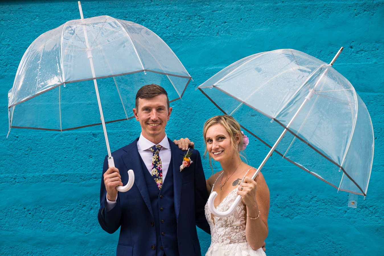 vibrant, fun image of the bride and groom holding clear umbrellas as they stand next to one another in front of a vibrant blue wall 