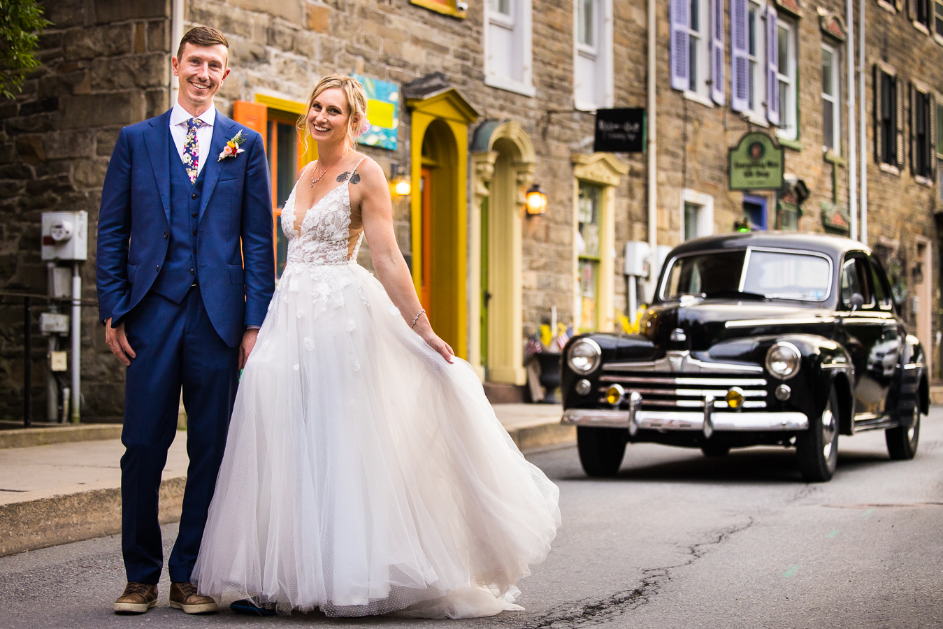 creative jim thorpe wedding photographer, lisa rhinehart, captures this image of the bride and groom holding hands and standing next to each other with a unique, old car in the background and vibrant colorful doors mixed in amongst the stone wall 