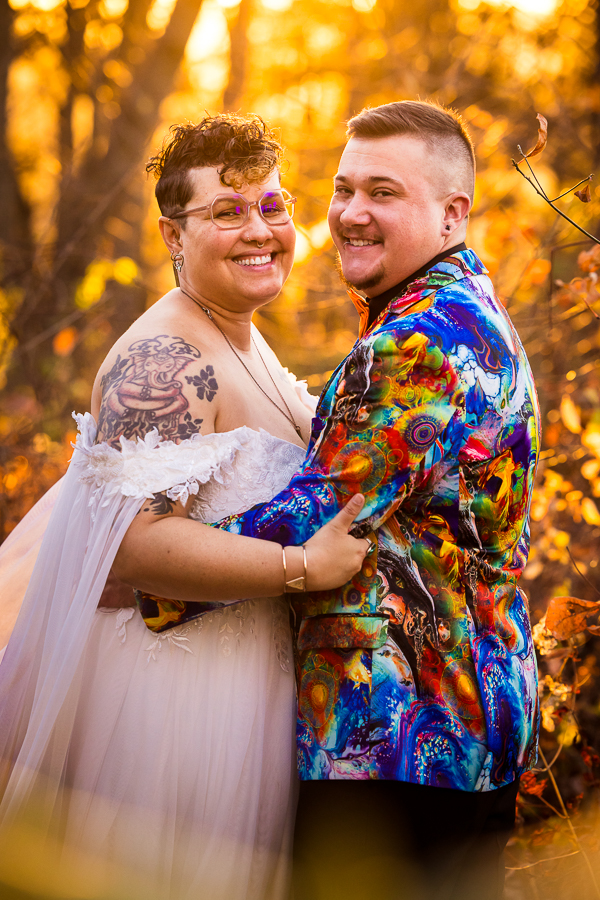 traditional portrait of the couple as they embrace one another and smile at the camera in their colorful wedding attire surrounded by the vibrant yellow glow from the sun and leaves around them 