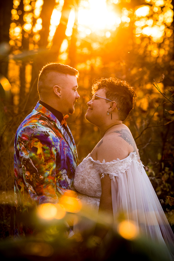 LGBTQ Wedding Photographer, lisa rhinehart, captures this creative and vibrant image of the couple as they stand in the woods smiling at each other surrounded by the trees and nature with the sun setting behind them 
