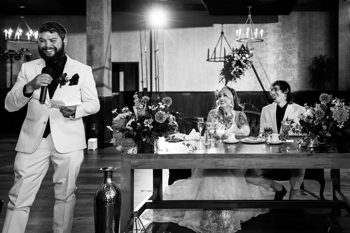 black and white image of the bride and groom sitting at their table as the best man gives his speech during the wedding reception in lancaster 