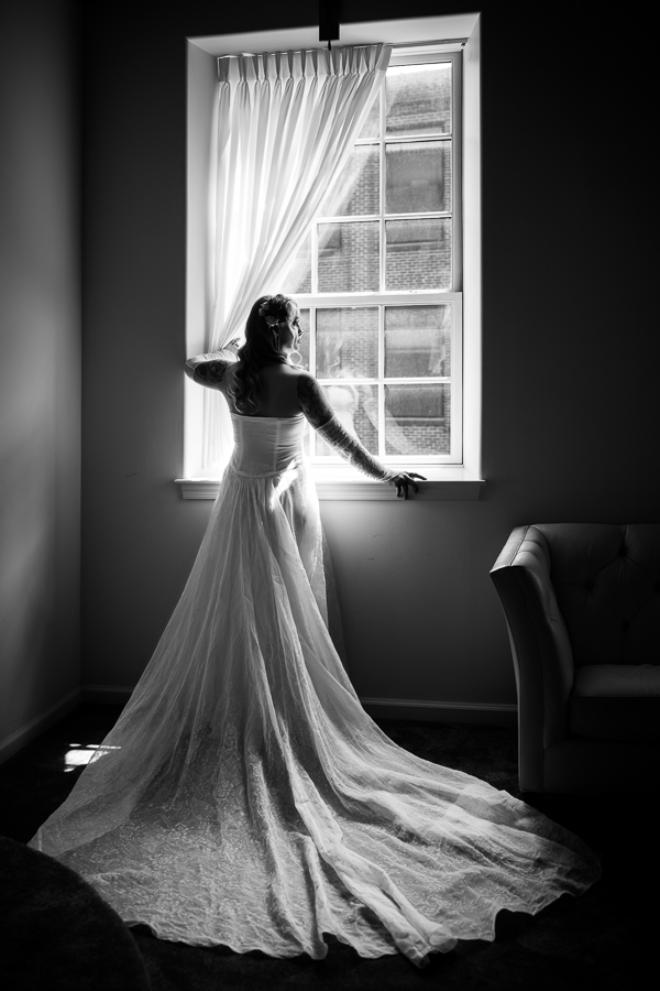 black and white image of the bride looking out the window holding back the white curtain captured by this Cork Factory Hotel Wedding Photographer