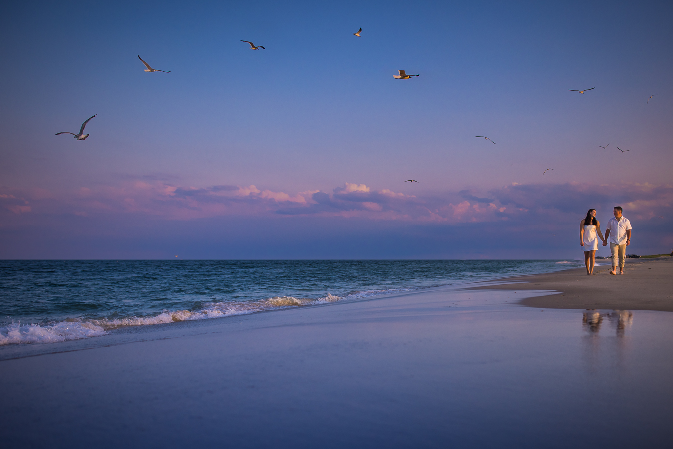 island beach state park photographer, lisa rhinehart, captures this vibrant, colorful image of the couple as they walk in the sand hand in hand looking each other by the ocean while the seagulls fly around them 