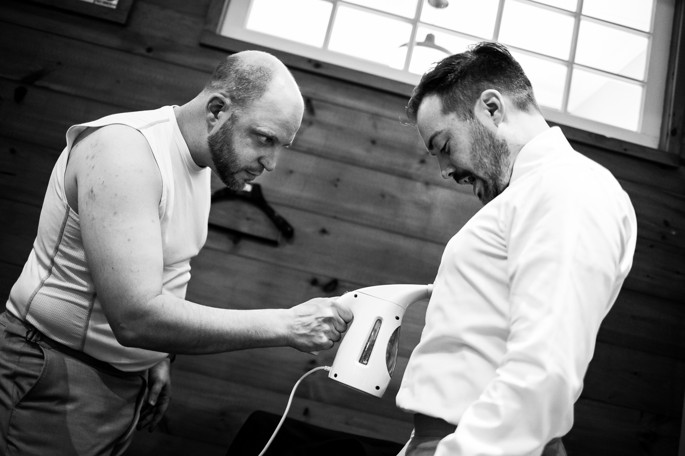 black and white image of the groom and a groomsmen steaming the grooms shirt as it is on him before the ceremony 