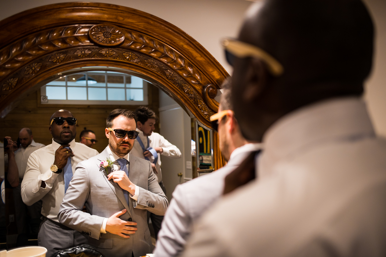 image of the groom getting ready with sunglasses on while he looks at himself in the mirror with one of his groomsmen behind him
