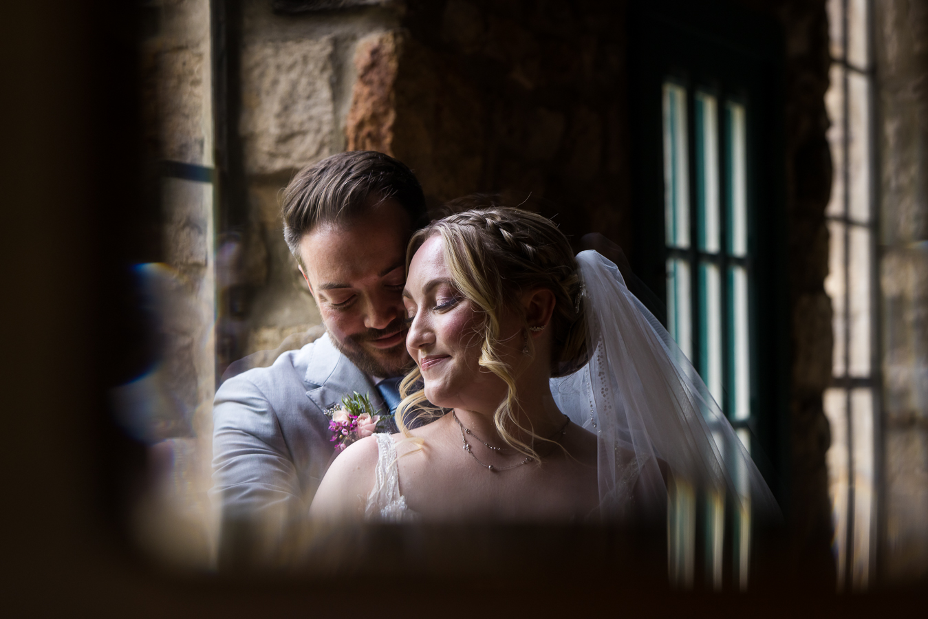 image of the bride and groom enbracing other another and smiling as they stand beside the big picture window captured through the glass on the piano by this creative Holly Hedge Estate Wedding Photographer