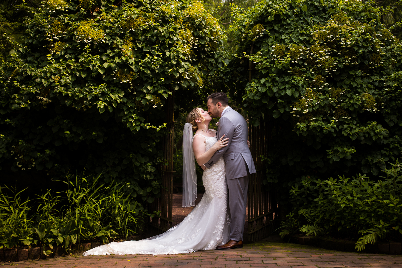 traditional image of the bride and groom hugging one another and kissing as they stand in front of the gate and are surrounded by lush greenery 