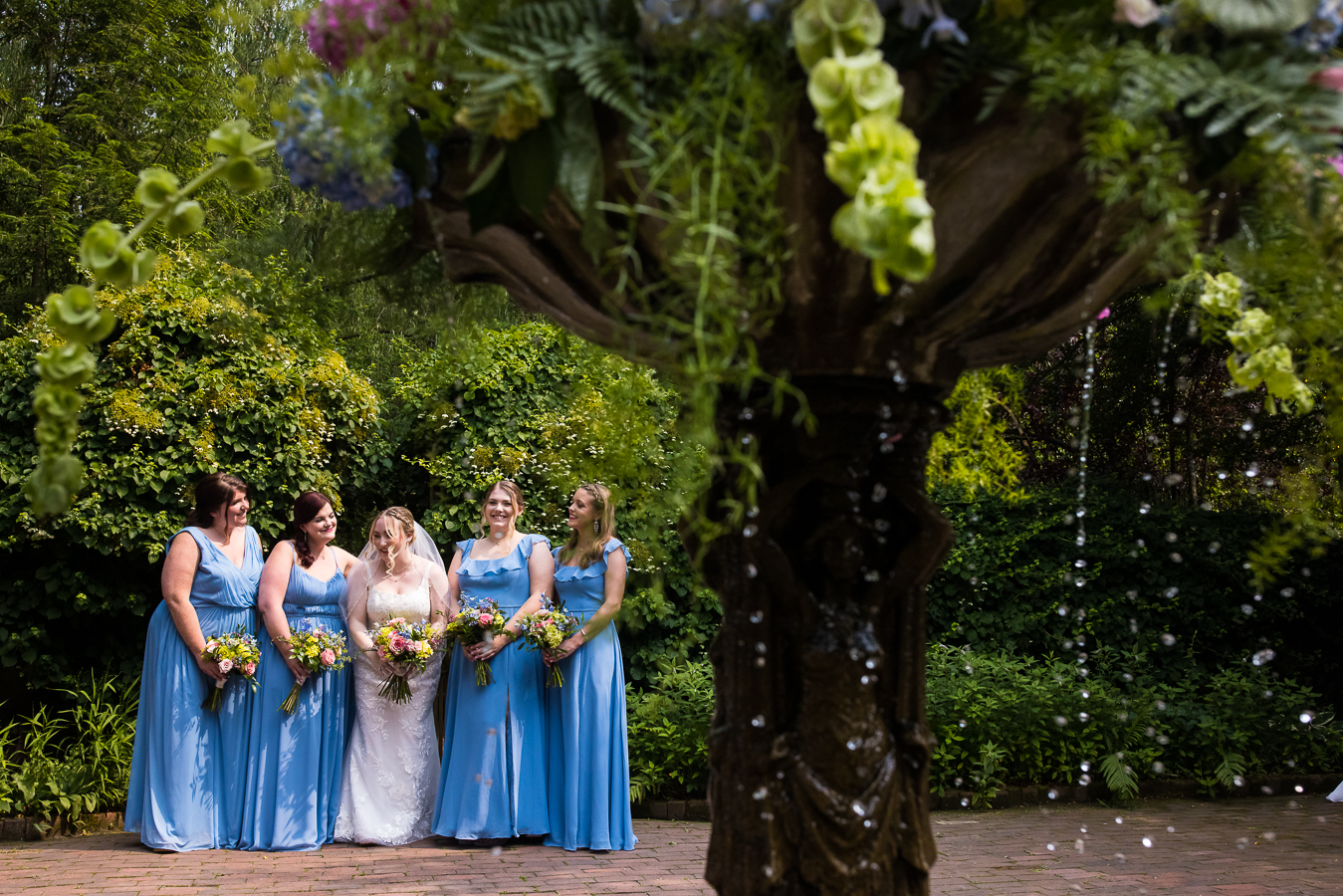candid photographer, lisa rhinehart, captures this image of the bride and her bridesmaids laughing and smiling at each other captured through the dripping outdoor water fountain 