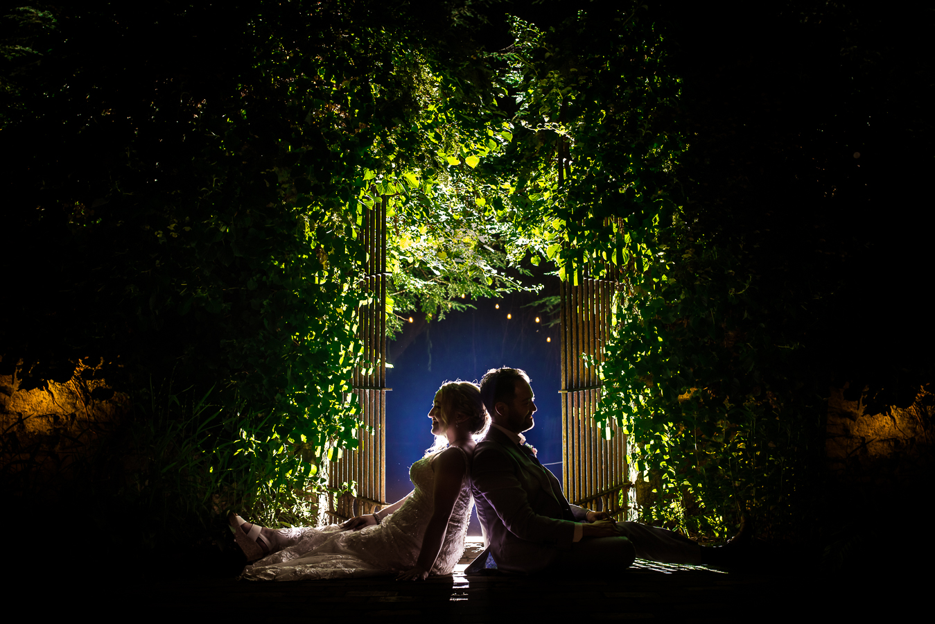 Lisa rhinehart, captures this unique, creative backlit image of the bride and groom sitting back to back surrounded by greenery in the archway 