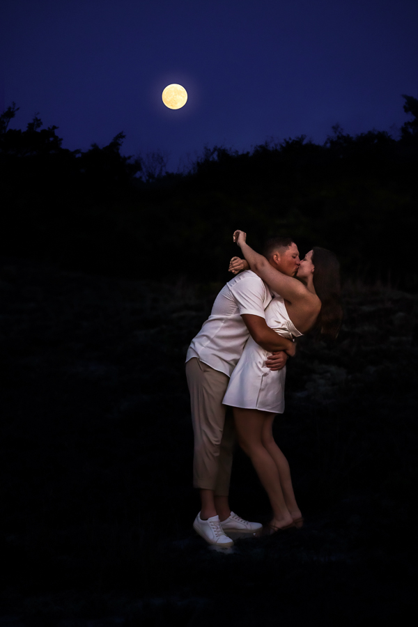 Image of the newly engaged couple as they kiss each other while the moon rises in the sky behind them during their NJ Shore Engagement