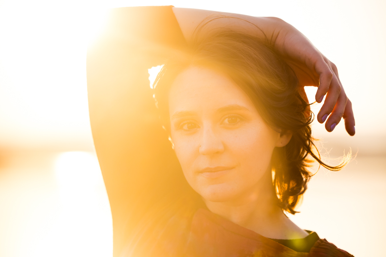 Image of this creative senior with her arm over her head and the bright vibrant yellow sun setting behind her during this riverfront portrait session
