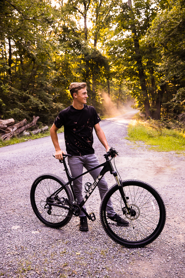 rhinehart photography, captures this portrait of this senior as he stands with his bike and looks off into the distance while the dust floats in the sunlight behind him on this riding trail in shippensburg pa 