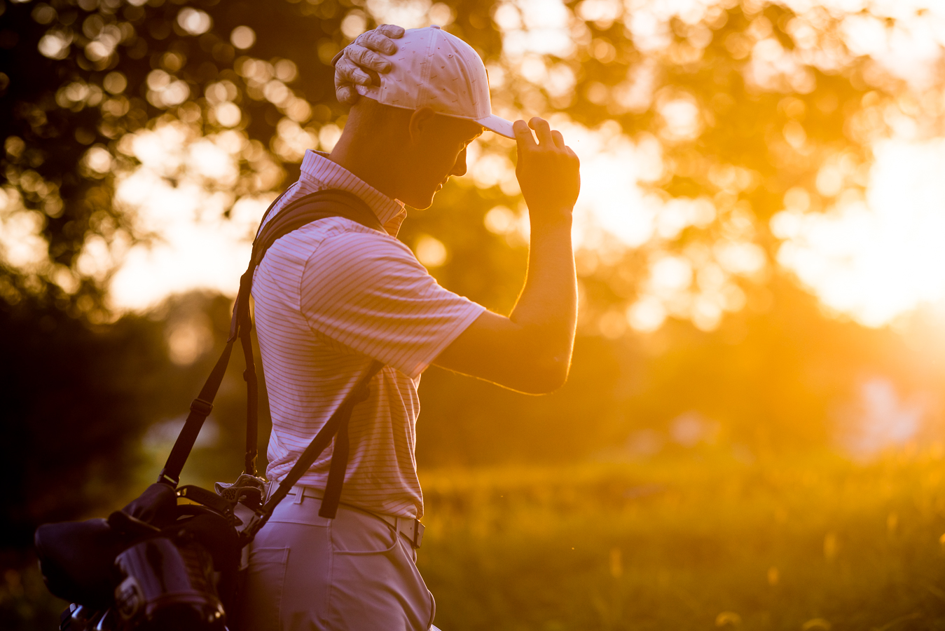 rhinehart photography, captures this vibrant, golden image of this senior as he puts his hat on while walking with his golf clubs at the Chambersburg country club 