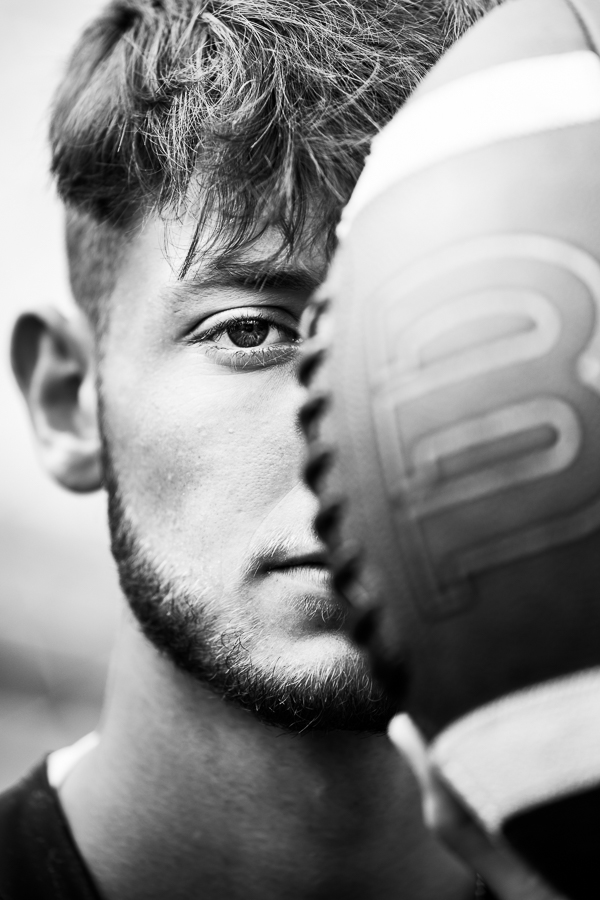 black and white image of a football player as he holds the ball in front of half his face while the other half peers out from behind the ball