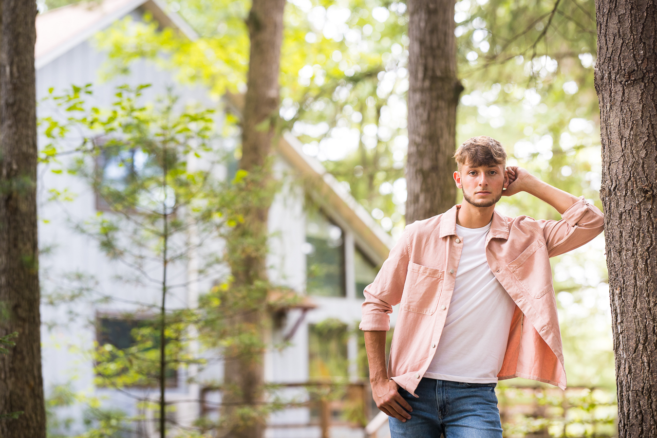 image of this male senior as he leans against the tree in the woods wearing a pink flannel top with a white t-shirt during this outdoor portrait session 