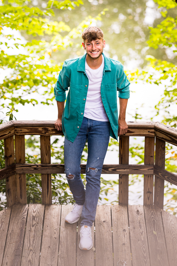 vibrant, colorful image of this male senior as he stands leaning against the outdoor wooden fence railing in the middle of the woods