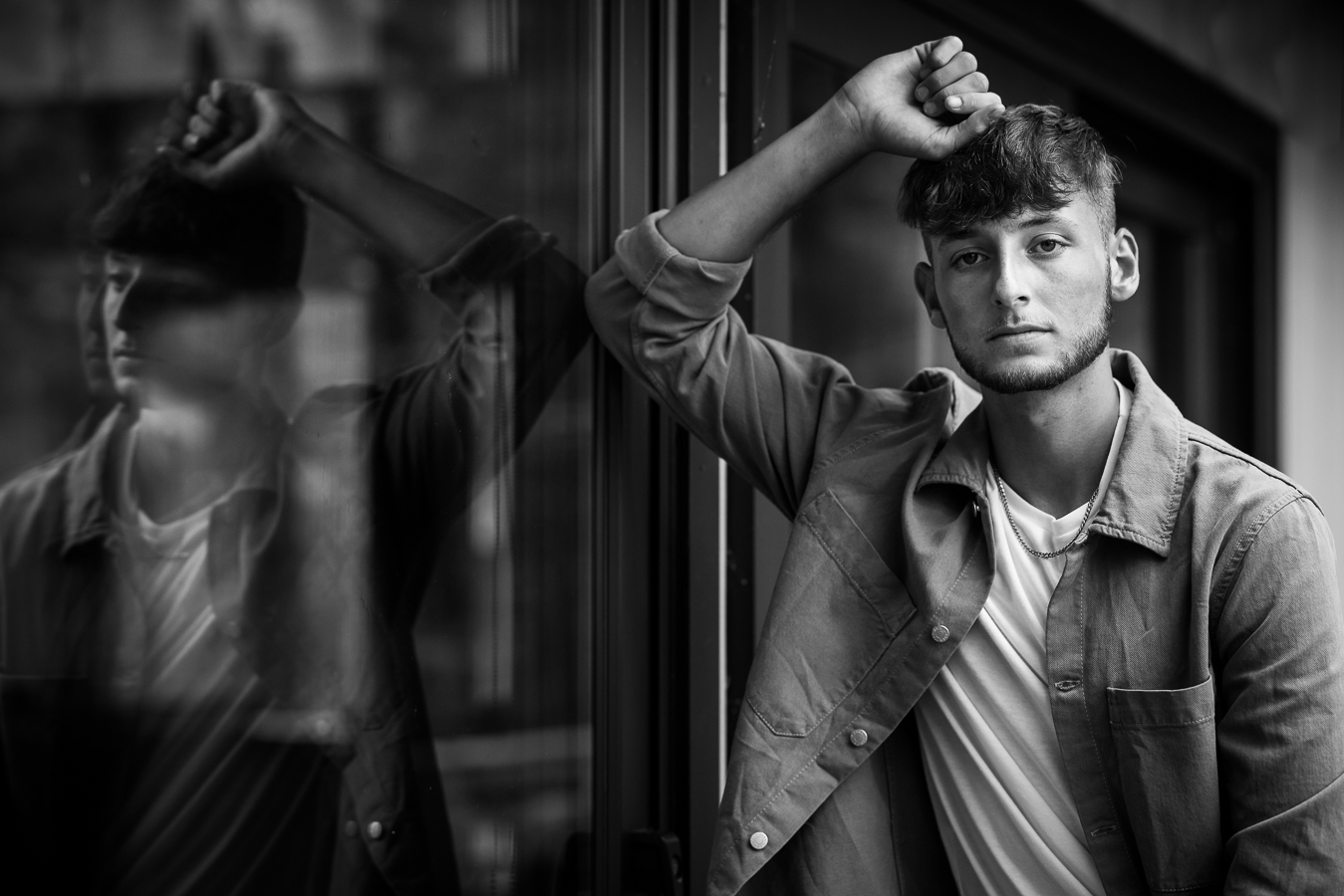 creative reflection in black and white of this senior as he leans up against the window and looks at the camera captured by High school Senior Portrait Photographer, rhinehart photography 