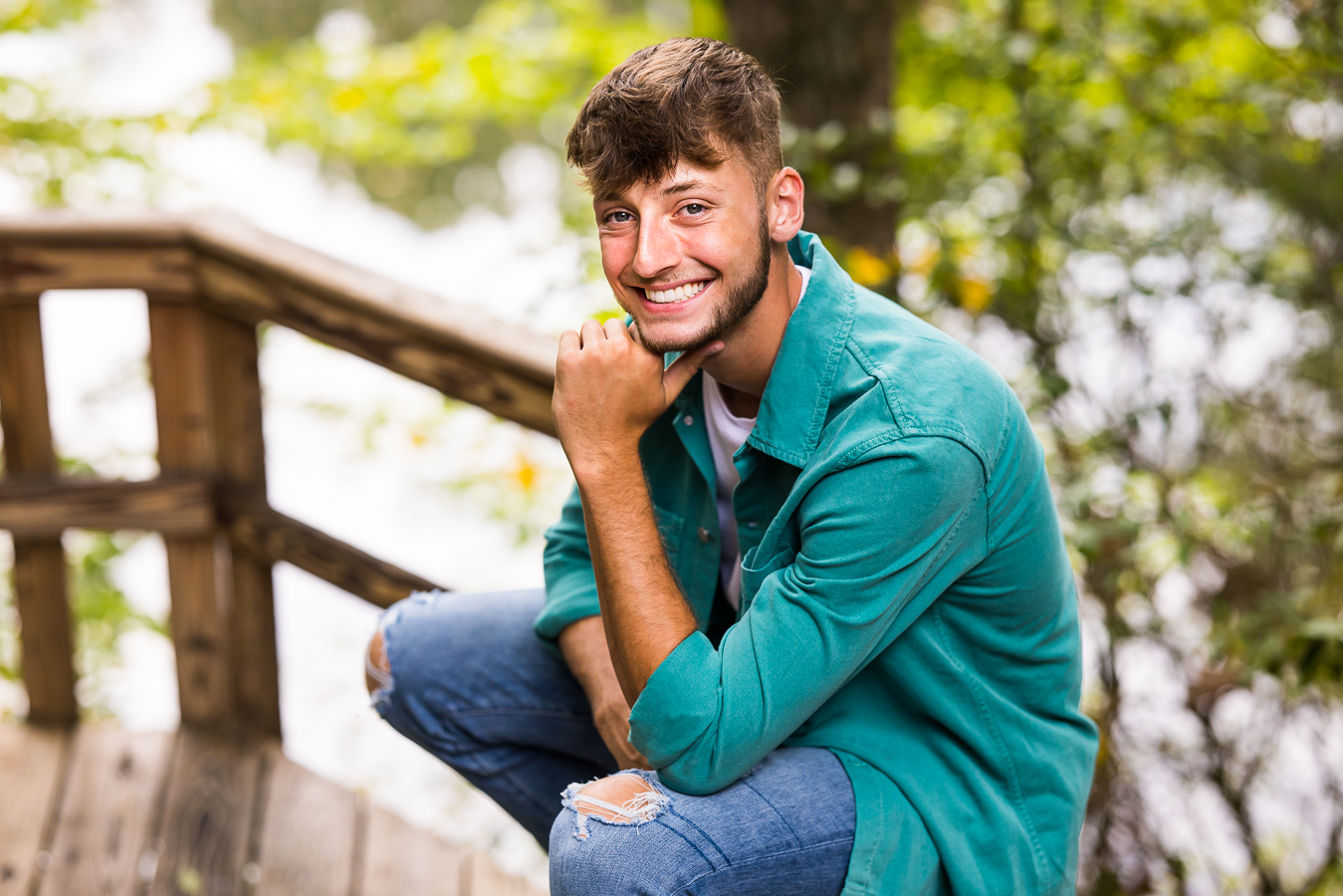 vibrant, colorful traditional portrait of this senior as he sits on the outdoor wooden railing with his arm propped on his knee as he smiles towards the camera in this outdoor session 