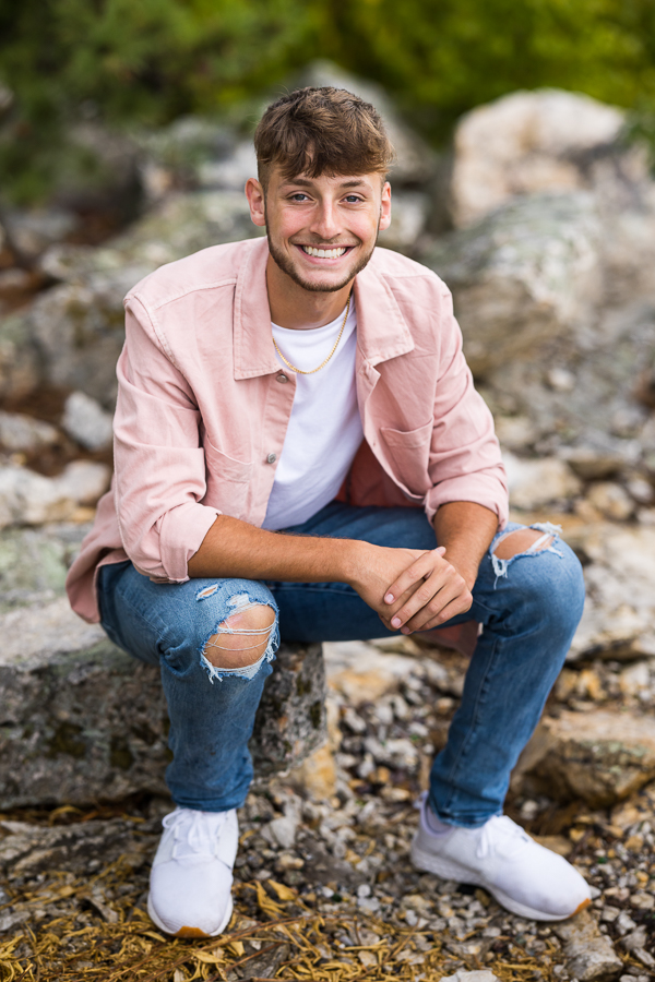 traditional portrait of this male senior as he sits on the rock overlook with his arms resting on his knees smiling at the camera in his pink flannel shirt