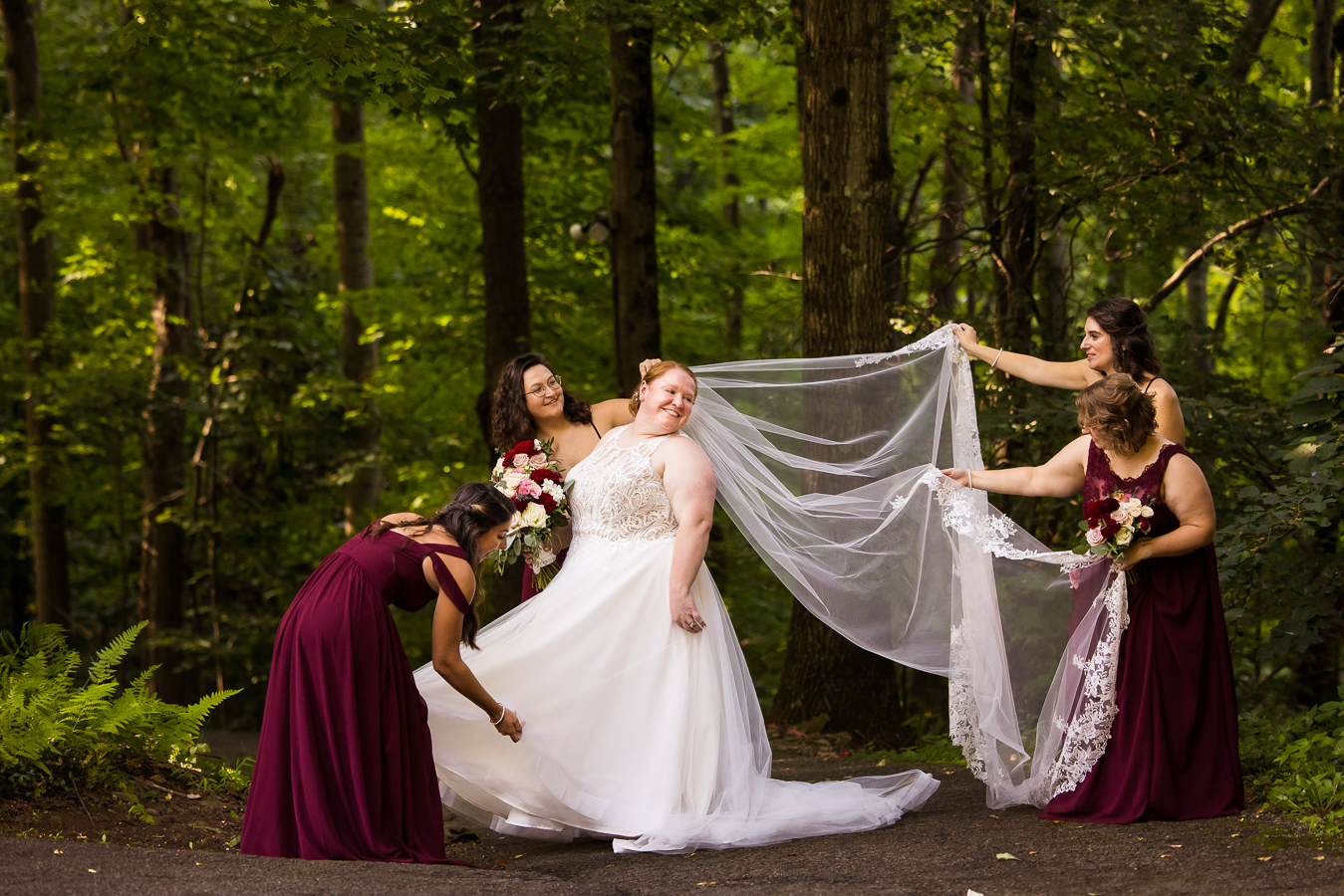 outdoor bridal party photographer, lisa rhinehart, captures this image of the bridesmaids dressed in maroon as they gather around the bride while laying out her veil and dress surrounded by the trees and mountains