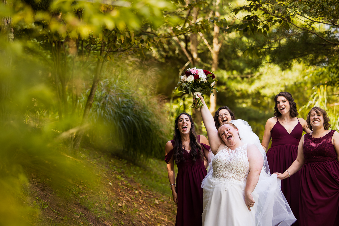 stahlstown outdoor photographer, rhinehart photography, captures this fun authentic image of the bride as she walks down the vibrant greenery outdoor pathway with her bridesmaids behind her as they smile and laugh together before the wedding ceremony at oak lodge