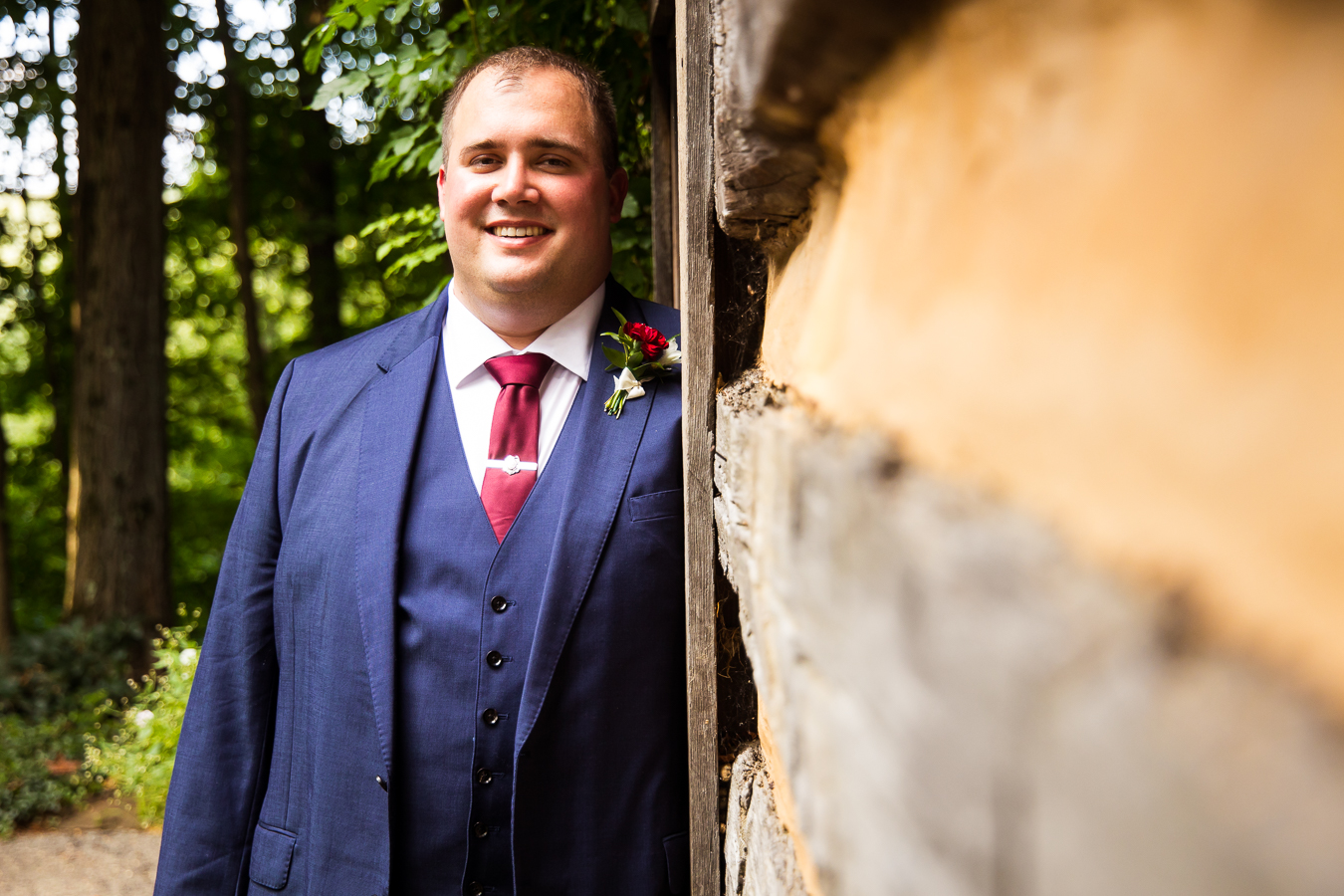 creative, unique image of the groom as he leans against the cabin walls and smiles at the camera in a traditional portrait prior to their wedding ceremony in stahlstown, pa 