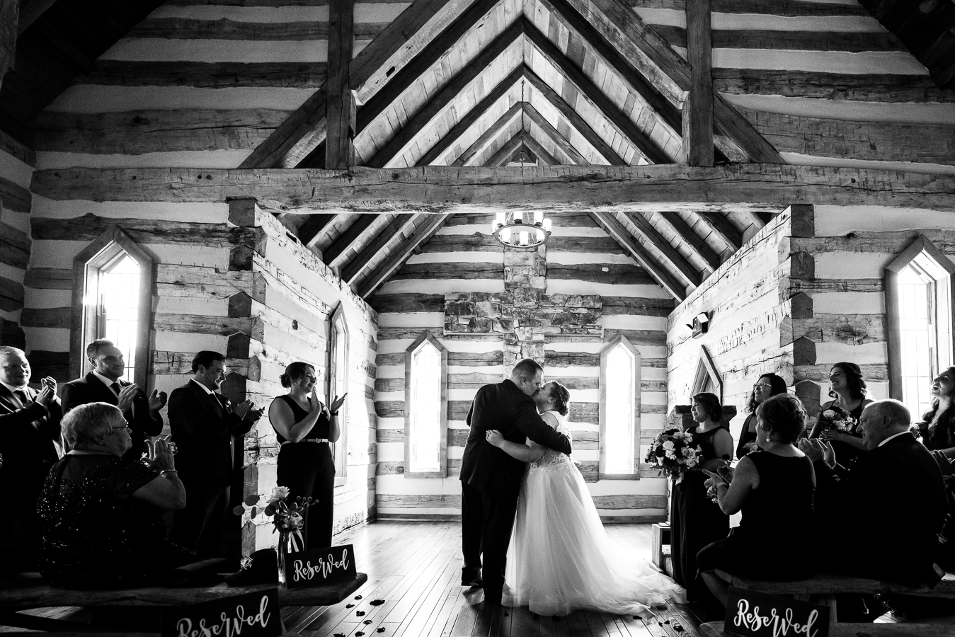 Oak Lodge Wedding Photographer, rhinehart photography, captures this black and white image of the bride and groom as they share their first kiss as husband and wife inside of this cozy log chapel surrounded by family and friends during their wedding ceremony in stahlstown pa at oak lodge