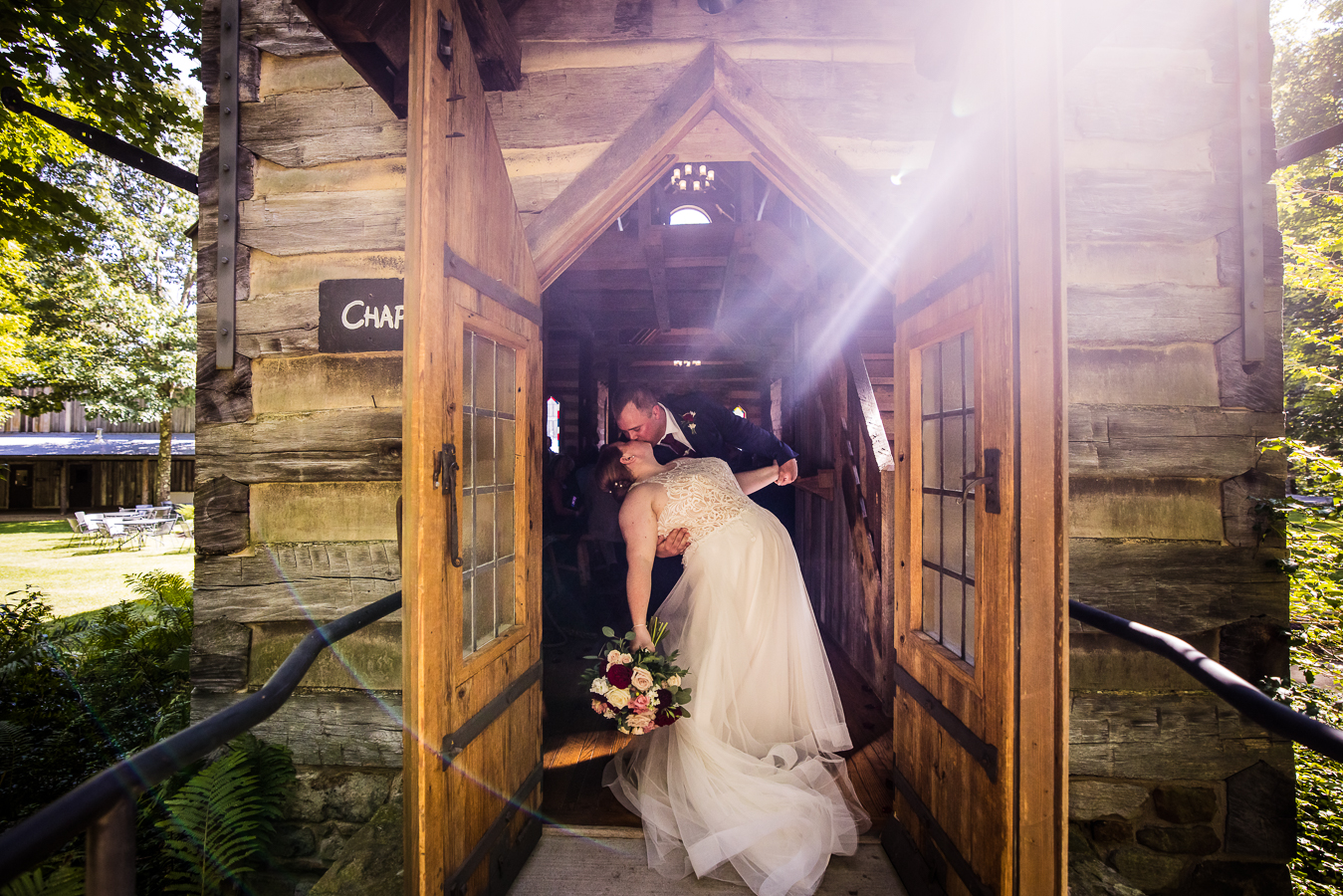 Oak Lodge Wedding Photographer, rhinehart photography, captures this romantic portrait of the bride and groom as they share a dip and a kiss at the entrance of the log chapel as a beam of light shines down on them during their kiss 