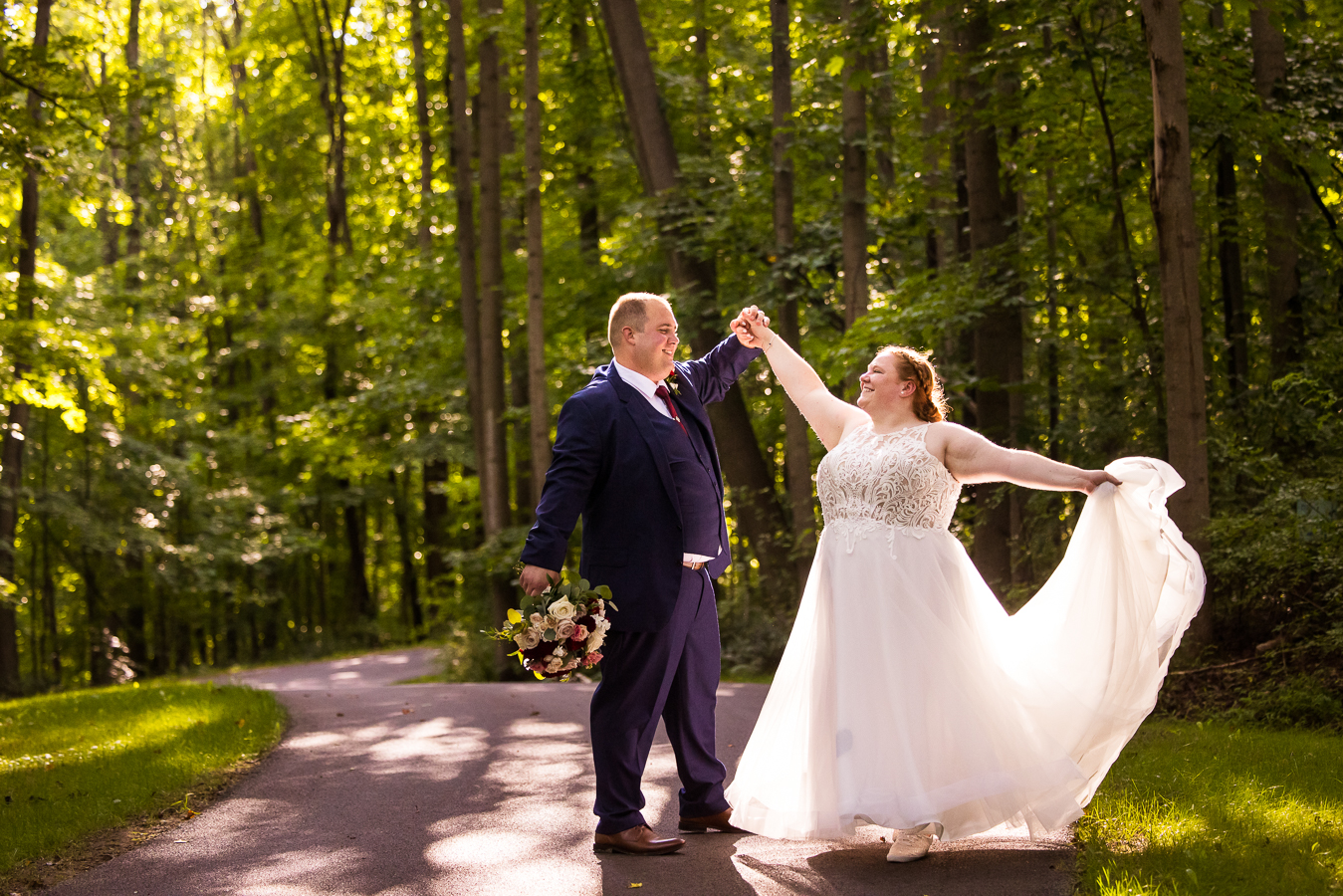 fun, vibrant image of the bride and groom as they walk down the pathway through the woods as the bride holds out her dress and spins during their outdoor fall wedding 