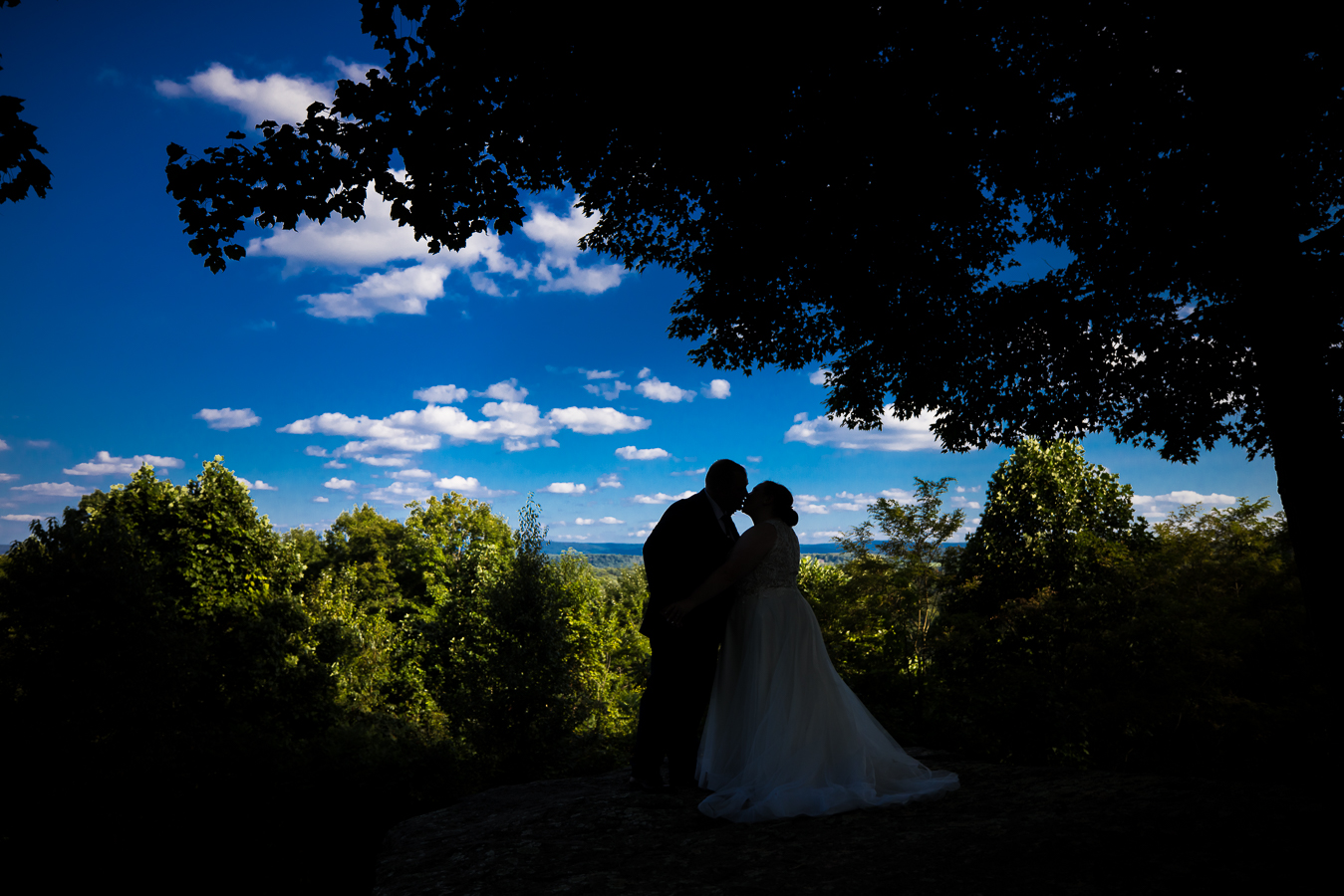 Oak Lodge Wedding Photographer, rhinehart photography, captures this vibrant image of the couple as they stand on a mountain overlook surrounded by trees and the vibrant blue sky during their outdoor wedding 