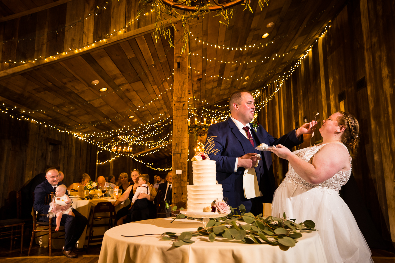barn wedding photographer, lisa rhinehart, captures this fun image of the groom feeding the bride some cake during their wedding reception with the gorgeous barn behind them decorated in the twinkle lights and greenery