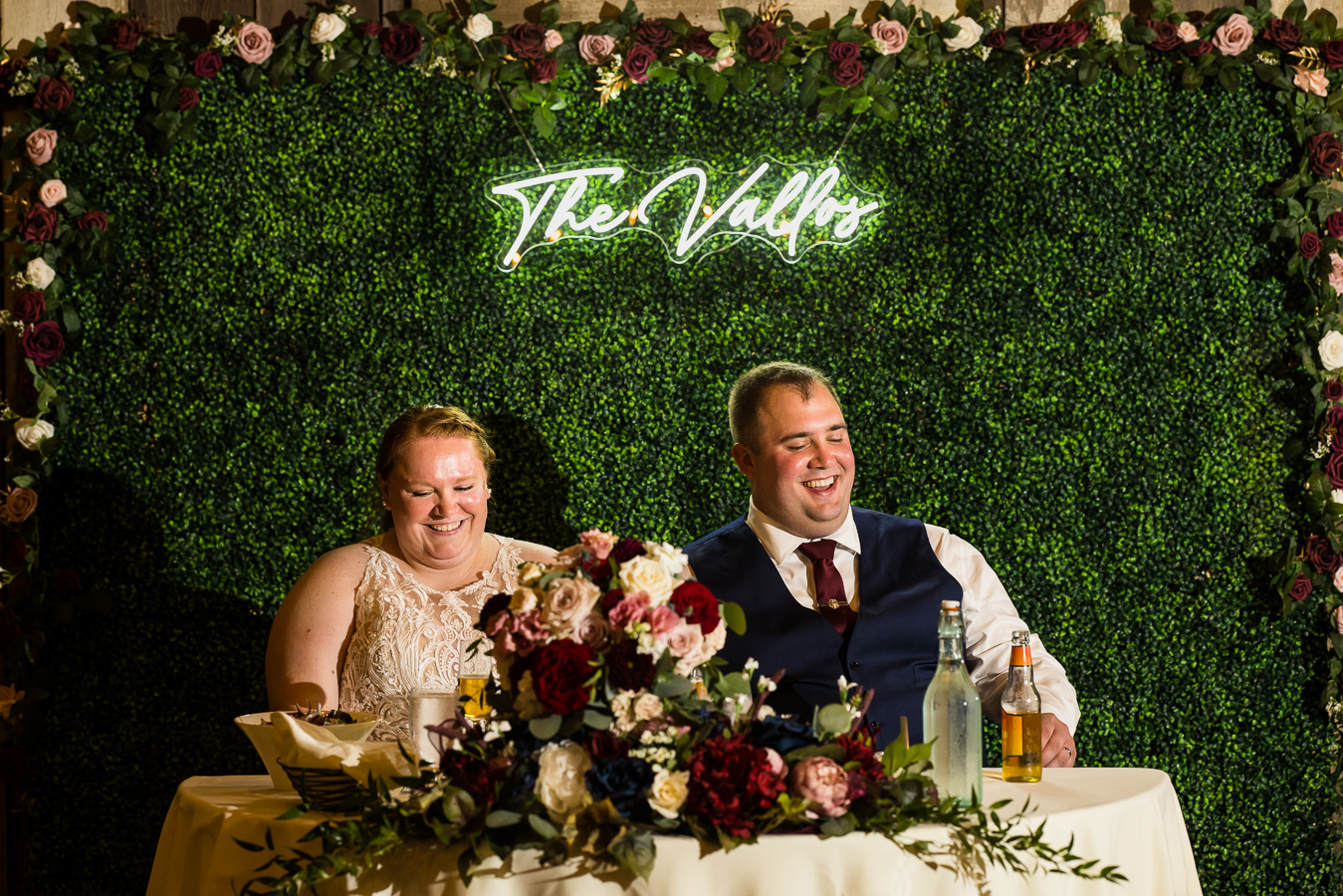 fun, unique greenery wall backdrop with their new last name on a neon sign behind the newlywed couple's sweetheart table