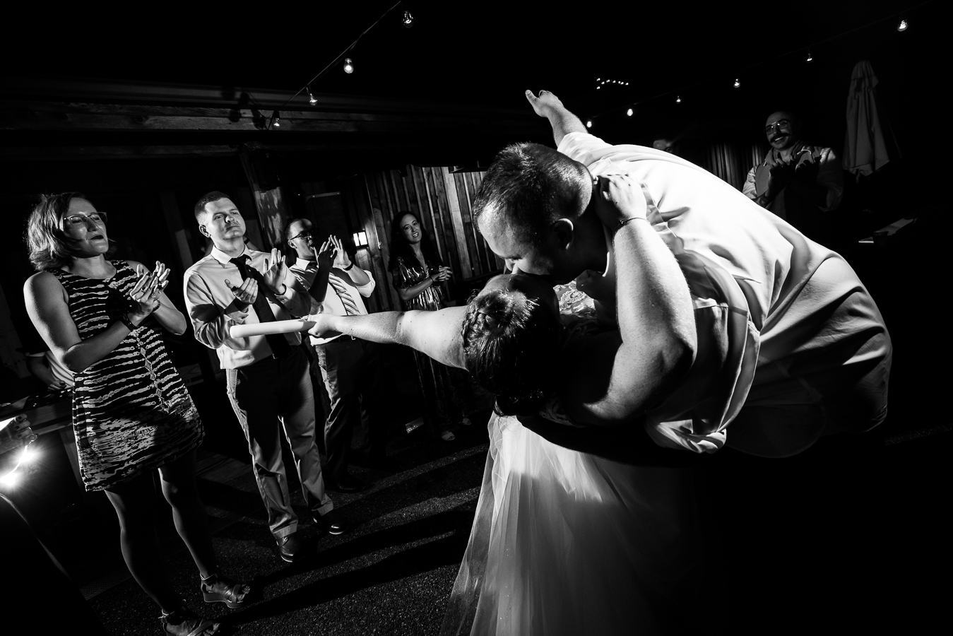 Oak Lodge Wedding Photographer, rhinehart photography, captures this black and white image of the bride and groom as they share and end of the night kiss while dipping as their family and friends gather around them at their outdoor wedding reception at oak lodge in stahlstown pa 