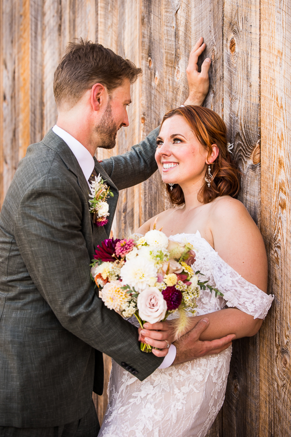 image of the couple as they lean against the rustic barn at alpine acres smiling at each other as they hold florals done by iron willow floral designs 