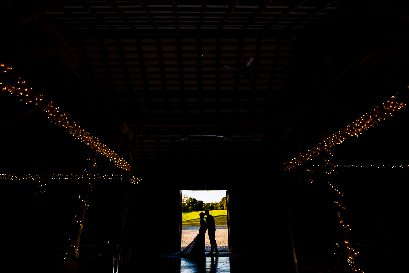 silhouetted image of the bride and groom as they stand and kiss in the doorway of the rustic barn with the lights on the beams showing through at alpine acres one of Central PA Wedding Venues located in warfordsburg pa 