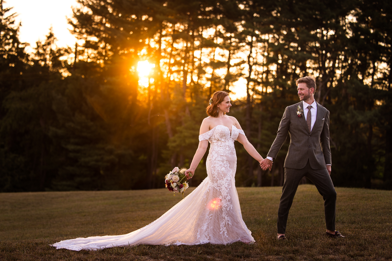 image of the bride and groom as they hold hands and walk across the open field as the golden sun is setting behind them peeking through the trees at this central pa wedding venues in warfordsburg, pa 