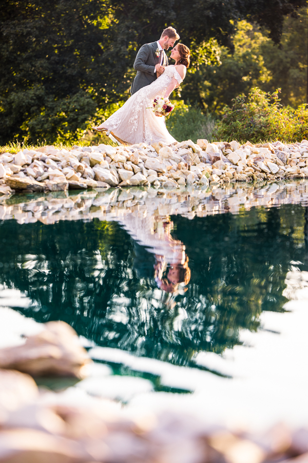 vibrant, colorful image of the bride and groom as they stand near the vibrant blue pond and lean backward as their reflection is captured in the pond during this creative branding photography session 