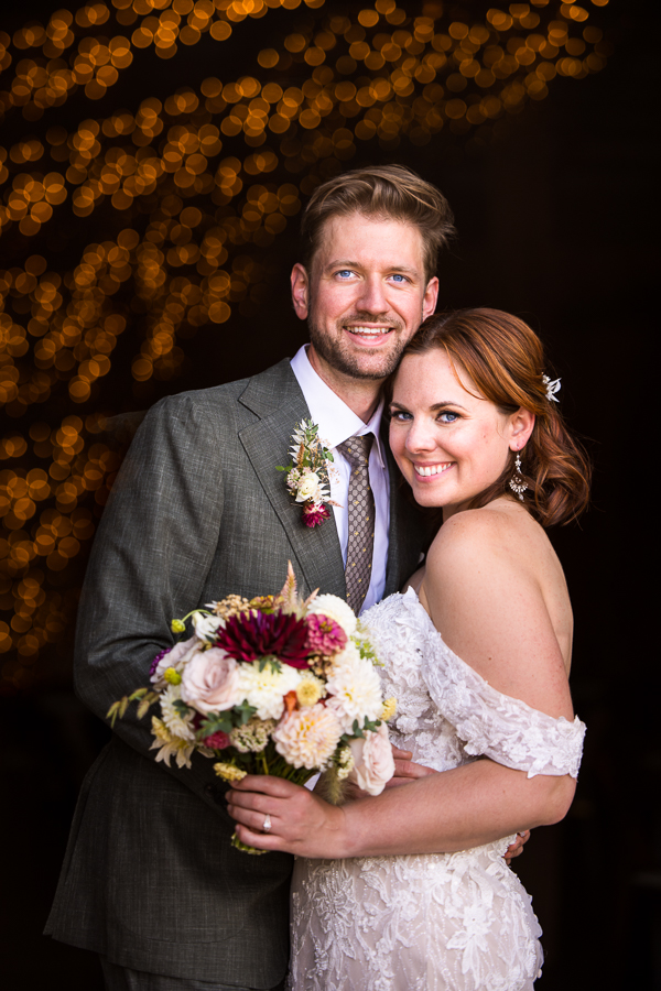 image of this bride and groom as they stand in the doorway of this rustic barn in a traditional hugging pose with the twinkle lights in the barn blurred out behind them 