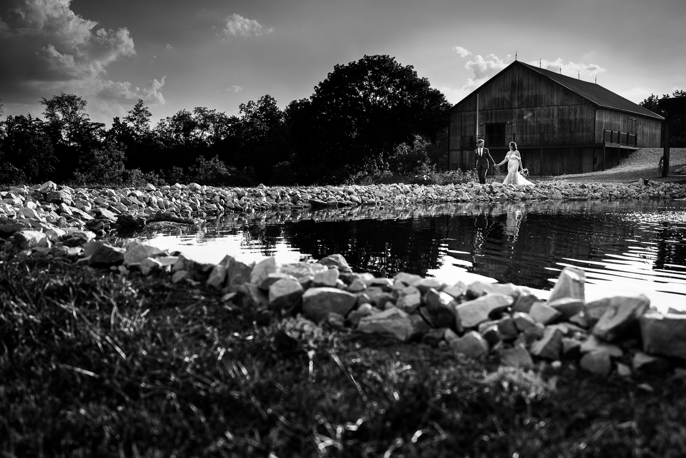 black and white image of the couple as they explore Alpine acres and walk beside the stunning pond with the barn in the background behind them during this branding photography session 