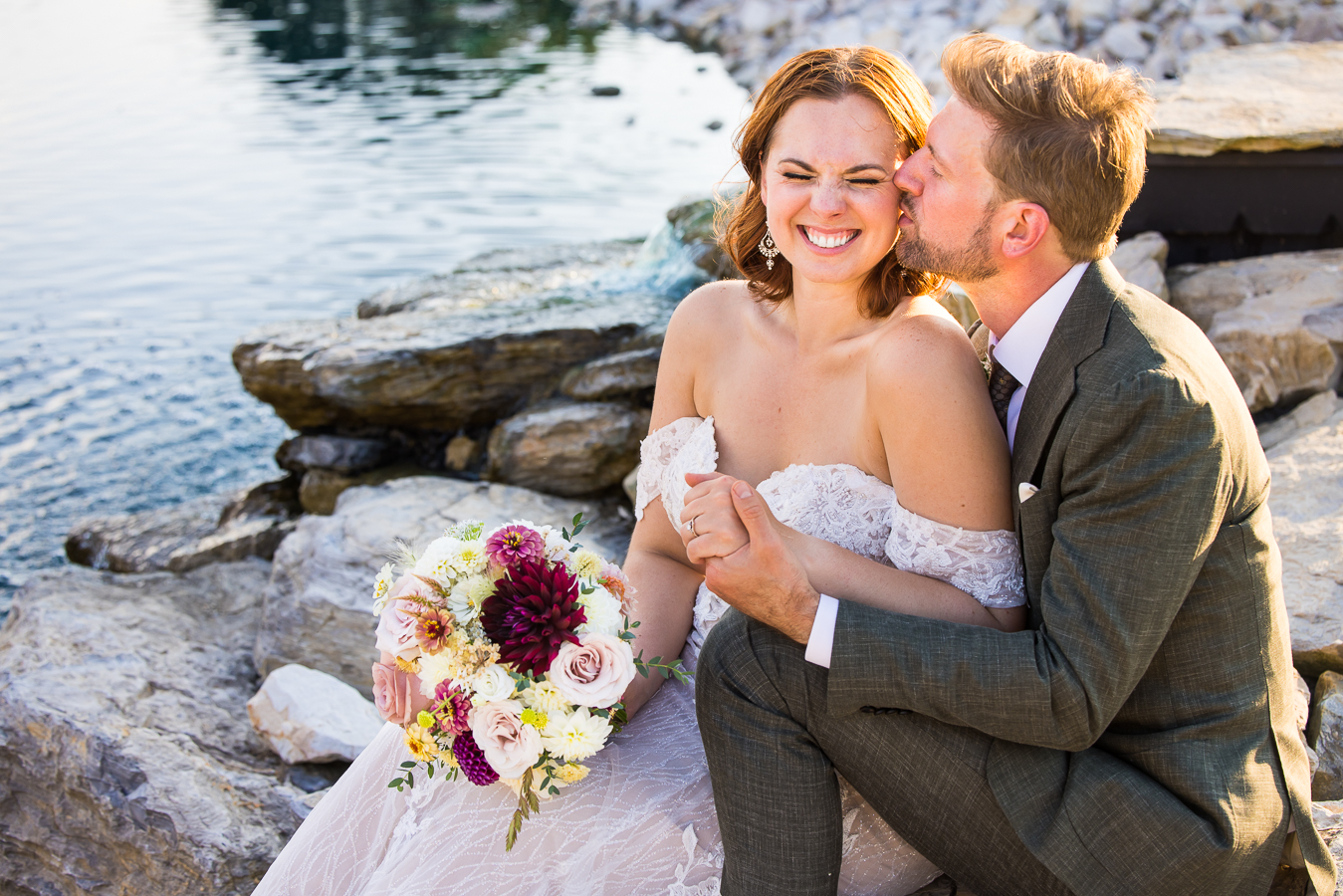 image of the bride and groom as they sit on the rocks near the vibrant blue pond and the groom kisses her on the cheek while they hold hands 
