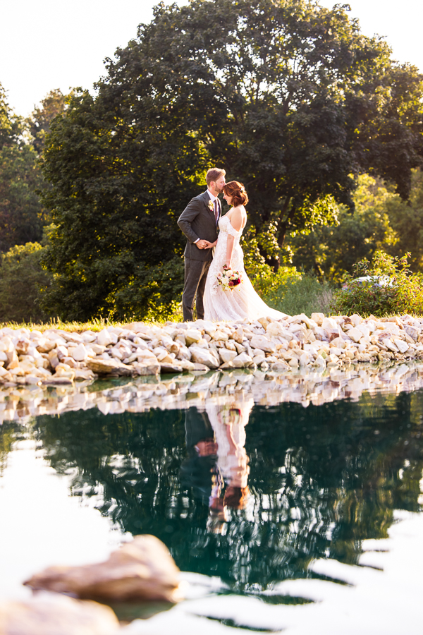 creative central pa wedding photographer, lisa rhinehart, captures this shot of the bride and groom as they hold hands and the groom kisses her forehead as they stand on the rocks near the pond with their reflection caught in the vibrant blue water during this branding photography session 