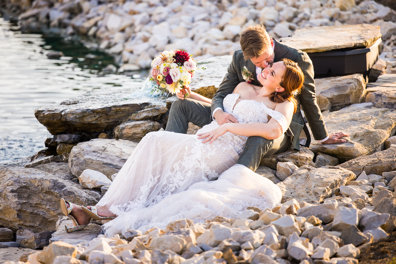 fun vibrant image of the bride and groom as he kisses her cheek and she laughs while they sit on the rocks beside the vibrant blue pond at alpine acres in warfordsburg pa 