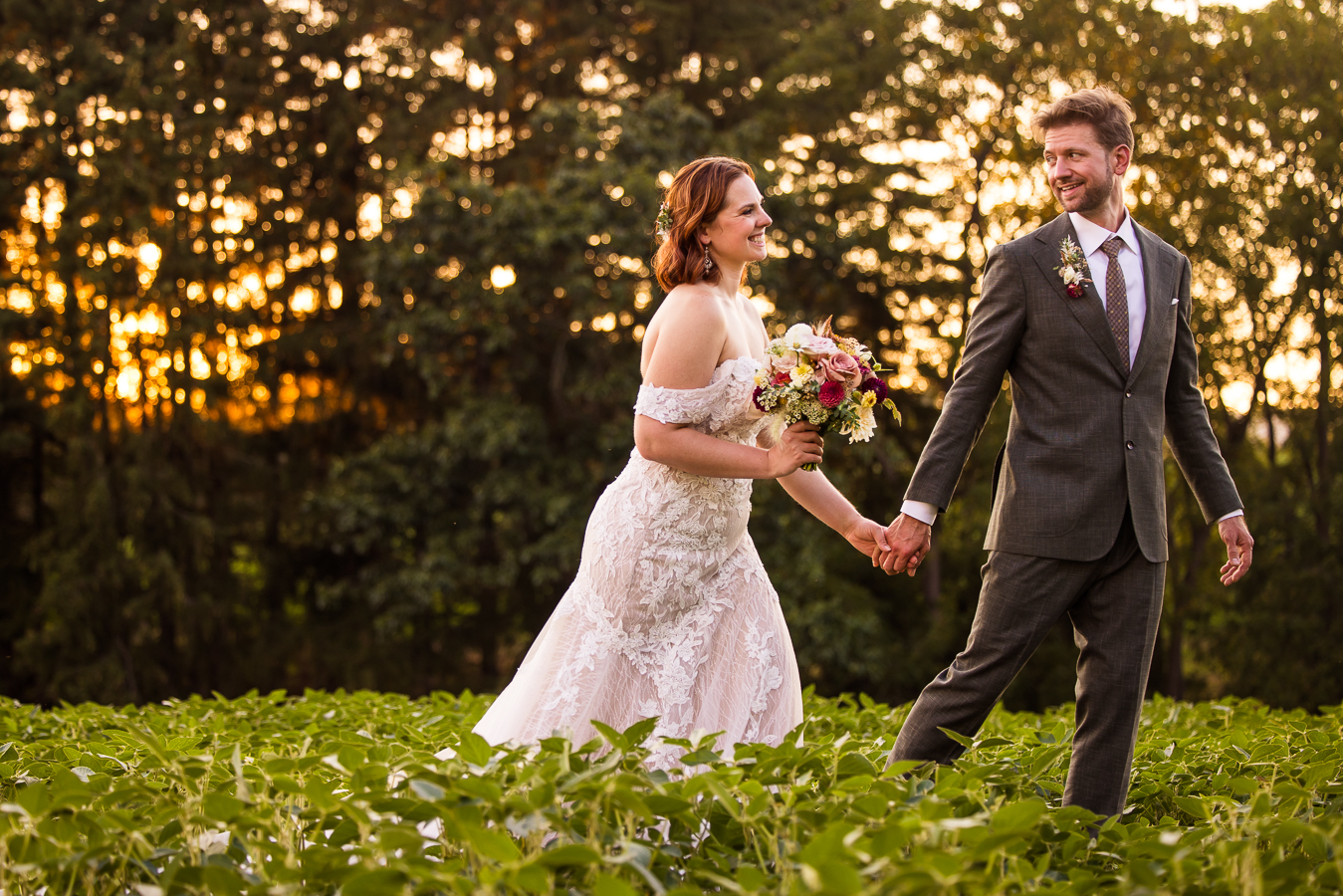 image of the bride and groom as they walk through the crops holding other another's hands and smiling as the sun sets behind them at alpine acres 